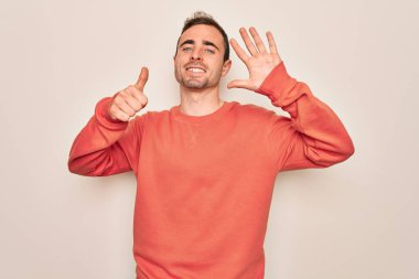 Mavi gözlü genç adam beyaz arka planda dikilip altı numarayı gösterirken kendinden emin ve mutlu bir şekilde gülümsüyor..