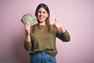 Dolarları elinde tutan genç ve güzel bir kadın pembe arka planda mutlu bir şekilde gülümsüyor tamam işareti yapıyor, parmaklarıyla başparmağını kaldırıyor, mükemmel bir işaret.