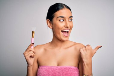 Genç, güzel esmer kadın duş aldıktan sonra havlu giyiyor kozmetik ürünleri kullanıyor spatula kullanıyor ve gülümseyen bir gülümsemeyle başparmağıyla yüzünü gösteriyor.