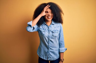 Sarıya boyanmış arka planda duran Afro saçlı güzel Afro-Amerikalı kadın. El hareketleriyle gülümsüyor, gözleriyle mutlu yüzüyle parmaklarının arasından bakıyor..