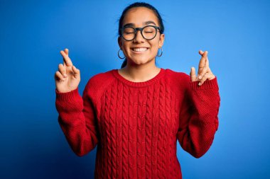 Genç ve güzel Asyalı bir kadın mavi arka plan üzerinde gözlük ve süveter takıyor umut ve gözler kapalı gülümseyerek el sallıyor. Şans ve batıl inanç..