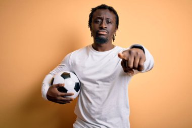 Genç Afrikalı Amerikalı oyuncu futbol topunu sarı zemin üzerinde tutarken kamerayı işaret ediyor ve sen, el işareti, ön taraftan pozitif ve kendinden emin bir jest.