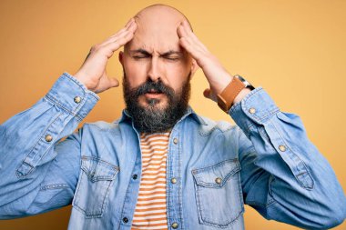 Sakallı, sıradan bir kot ceket giyen ve baş ağrısı ve ağrı ve migren yüzünden stresli olan çizgili tişörtlü yakışıklı kel bir adam. Ellerini başının üstüne koy..