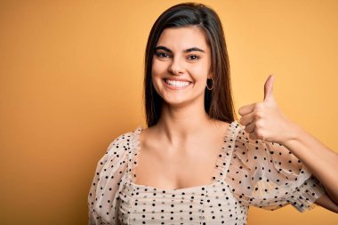 Sarı arka plan üzerinde sıradan tişörtler giyen genç, güzel esmer bir kadın mutlu bir el hareketi yapıyor. Kameraya bakıp başarıyı gösteren ifade onaylanıyor.