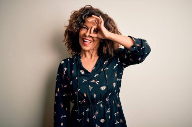 Orta yaşlı, güzel bir kadın, sıradan bir elbise giymiş, izole edilmiş beyaz arka planda duruyor, el hareketleriyle gülümsüyor, gözleriyle mutlu yüzle parmaklarının arasından bakıyor..