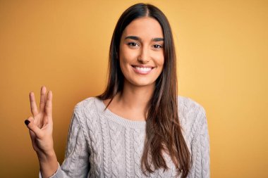 Sarı arka planda beyaz süveter giyen genç, güzel esmer kadın üç numaralı parmaklarını gösterirken kendinden emin ve mutlu bir şekilde gülümsüyor..
