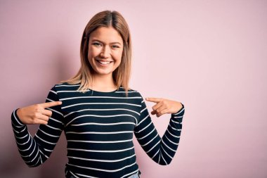 Pembe izole edilmiş arka plan üzerine sıradan çizgili süveter giyen genç, güzel sarışın kadın yüzünde bir gülümsemeyle, gururlu ve mutlu parmaklarıyla kendini işaret ediyor..