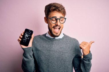 Sakallı, gözlüklü, kırık ve çatırdayan akıllı telefonlu genç adam gülümseyerek baş parmağıyla yüzünü gösteriyor.