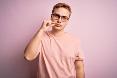 Genç, kızıl saçlı, sıradan bir tişört giyen, izole edilmiş pembe arka plan ağzının üzerinde duran dudakları parmaklarıyla kapanmış genç bir adam. Gizli ve sessiz, tabu konuşması