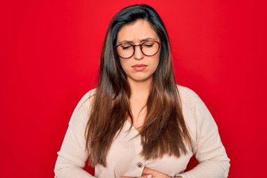 Kırmızı izole edilmiş arka planda duran gözlük takan genç İspanyol kadın midesi ağrıyan, midesi ağrıyan ve hazımsızlık çeken hasta. Ache kavramı.