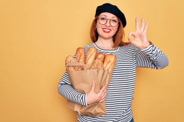 Sarı arka planda ekmek taşıyan, bereli genç bir kadın. Tamam işareti yapıyor, mükemmel bir sembol.