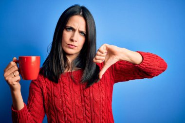 Mavi gözlü genç bir kadın izole edilmiş arka planda dikilmiş bir fincan kahve içiyor. Kızgın bir yüzü var. Eksi işareti var.