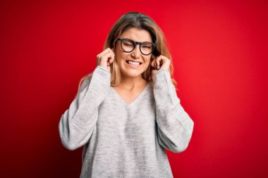 Genç, sarışın, süveter ve gözlük takan, izole edilmiş kırmızı arka plan üzerine yüksek sesli müzik sesi için parmaklarıyla kulaklarını kapatan güzel bir kadın. Sağır kavramı.