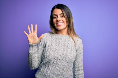 İzole edilmiş mor arka plan üzerinde duran sıradan bir süveter giyen genç ve güzel bir kadın beş numaralı parmaklarıyla işaret ederken kendinden emin ve mutlu gülümsüyor..