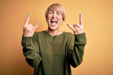 Modern kısa saçlı, sarı arka planda sıradan süveter giyen, çılgın bir ifadeyle elleri havada rock sembolü yapan genç sarışın kadın. Müzik yıldızı. Ağır müzik konsepti.