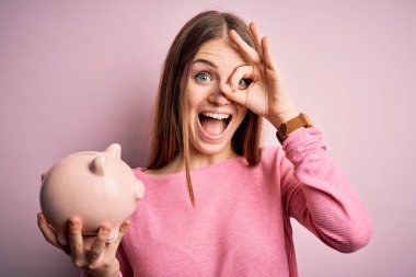 Genç, güzel, kızıl saçlı bir kadın kumbarayı izole edilmiş pembe arka planda tutuyor. Gülümsüyor, gülümsüyor.