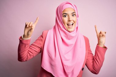 İslami tesettür giyen genç ve güzel bir kız pembe arka planda durmuş şaşkın şaşkın gülüyor ve parmaklarıyla işaret ediyor ve kollarını kaldırıyordu..