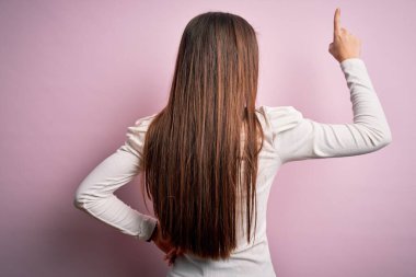 Mavi gözlü genç bir kadın pembe arka plan üzerine günlük beyaz tişörtler giyip parmak eliyle öne doğru poz veriyor.