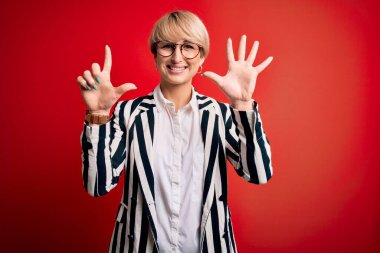 Sarışın, kısa saçlı, gözlüklü, kırmızı arka planda çizgili ceketli, kendine güvenen ve mutlu bir şekilde 7 numarayı gösteren iş kadını..