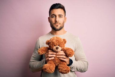 Genç yakışıklı adam elinde oyuncak ayıyla pembe arka planda dikiliyor. Kendine güvenen bir yüz ifadesi var ve ciddi düşünüyor.