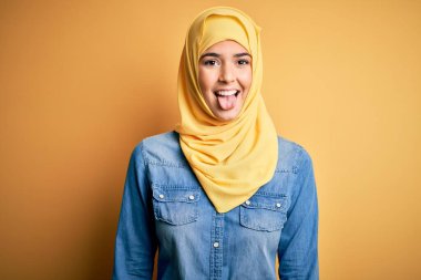 Genç, güzel bir kız, izole edilmiş sarı arka planda duran Müslüman başörtüsü takıyor ve komik bir ifadeyle dilini dışarı çıkarıyor. Duygu kavramı.