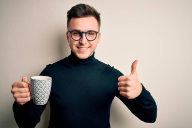 Genç, beyaz, gözlüklü bir adam bir fincan sıcak kahve içiyor. Kocaman gülümsüyor. İşaret veriyor.