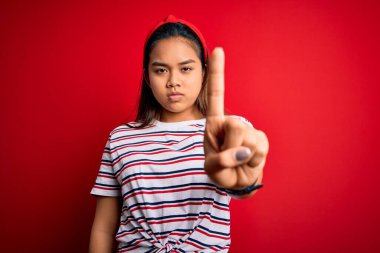 Genç, güzel Asyalı bir kız izole edilmiş kırmızı arka plan üzerine sıradan çizgili tişört giyiyor. Parmağını kaldırmış ve kızgın bir ifade sergiliyor.