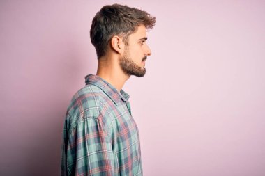 Sakallı, rahat gömlekli genç bir adam pembe arka planda dikiliyor, yan yana bakıyor, rahat poz veriyor, kendine güvenen bir gülümsemesi var..