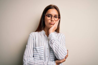 Genç, güzel esmer kadın beyaz arka plan üzerine günlük gömlek ve gözlük takıyor. Kollarını kavuşturup depresyondan sıkılmış ve yorgun görünüyor..