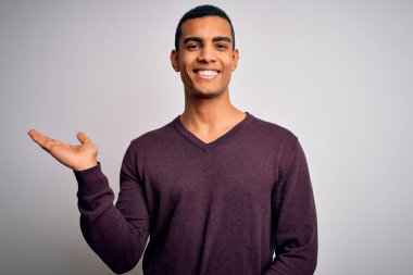 Beyaz arka plan üzerine gündelik süveter giyen genç ve yakışıklı Afro-Amerikan adam gülümsüyor ve avuç içi kameraya bakıyor..