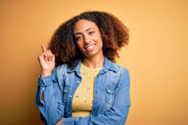 Afro saçlı, sarı arka planda sıradan bir kot gömlek giyen, yüzünde kocaman bir gülümseme olan, eli ve parmağı kameraya bakan Afro Amerikalı genç bir kadın..