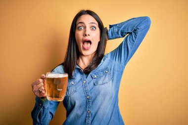 Mavi gözlü genç bir kadın, izole edilmiş sarı arka planda duran bira kavanozunu içiyor. Çılgına dönmüş, elleri başında, korkmuş ve ağzı açık bir şekilde şok geçirmekten korkan bir kadın.