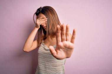 Genç, güzel esmer kız izole edilmiş pembe arka plan üzerine günlük çizgili tişört giyiyor gözleri ellerle örtüyor ve üzgün ve korku ifadeleriyle dur hareketi yapıyor. Utanç verici ve olumsuz bir kavram..