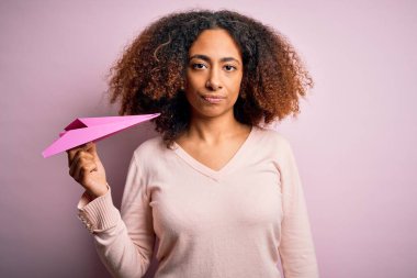 Afro saçlı, pembe arka planda kağıt uçakla gezen Afro-Amerikalı genç bir kadın.