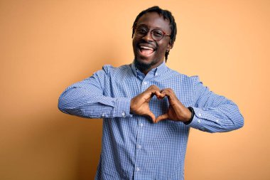Sarı arka planda gömlek ve gözlük takan yakışıklı Afro-Amerikan bir adam. Sevgiyle gülümsüyor, elleriyle kalp sembolü yapıyor. Romantik konsept.