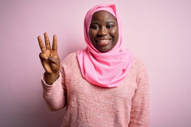 Genç Afro-Amerikan artı cüsseli kadın soyutlanmış pembe arka plan üzerine Müslüman başörtüsü takıyor ve üç numarayı işaret ederek kendinden emin ve mutlu bir şekilde gülümsüyor..
