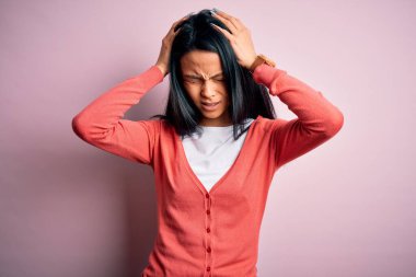 Genç, güzel, Çinli bir kadın pembe arka plan üzerine günlük süveter giyiyor. Ağrı ve migren yüzünden baş ağrısı ve stresten muzdarip. Ellerini başının üstüne koy..