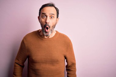 Orta yaşlı, beyaz, günlük kahverengi süveter giyen, pembe arka planda duran, korku, korku ve heyecan dolu bir yüz ifadesinden korkan ve şok olan bir adam..