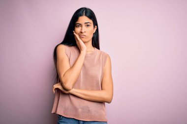Pembe arka planda günlük süveter giyen genç, güzel İspanyol moda kadını kollarını kavuşturarak depresyondan sıkılmış ve yorgun görünüyor..