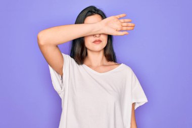 Genç, güzel, esmer bir kadın mor arka plan üzerine gündelik beyaz tişört giyip gözlerini kollarıyla kapatıyor, ciddi ve üzgün görünüyor. Görme, saklama ve reddetme konsepti