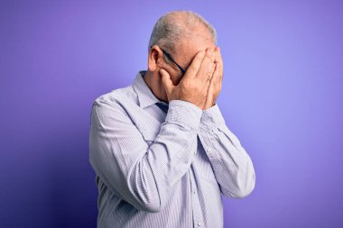 Orta yaşlı, beyaz, çizgili gömlek ve mor arka planda gözlük takan yakışıklı bir adam. Ağlarken yüzünü ellerle kapatan üzgün bir ifade. Depresyon kavramı.