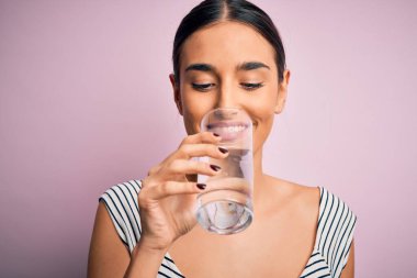 Genç ve güzel esmer kadın mutlu ve kendinden emin gülümsüyor. Yüzünde gülümsemeyle dikilip, izole edilmiş pembe arka planda ferahlamak için bir bardak sağlıklı su içiyorum.