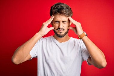 Sakallı genç yakışıklı bir adam kırmızı arka planda dikiliyor. Ağrı ve migren yüzünden baş ağrısı çekiyor. Ellerini başının üstüne koy..