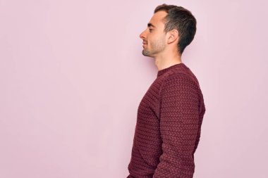 Mavi gözlü, sıradan bir süveter giyen, pembe arka planda duran, rahat bir görünüm takınan, kendine güvenen ve gülümseyen genç yakışıklı bir adam..