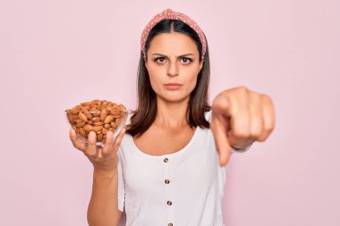 Genç, güzel esmer kadın elinde sağlıklı bademlerle pembe arka planda kaseyle kamerayı ve seni işaret ediyor. Kendine güveni tam bir jest.