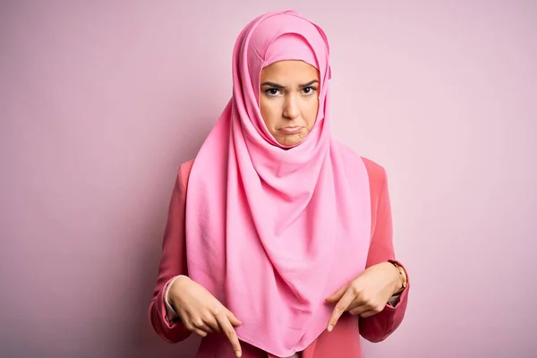若いです美しいです女の子身に着けていますイスラム教徒のヒジャーブ立って上の隔離されたピンクの背景を指します悲しいと動揺 指で方向を示します 不幸で落ち込んで — ストック写真