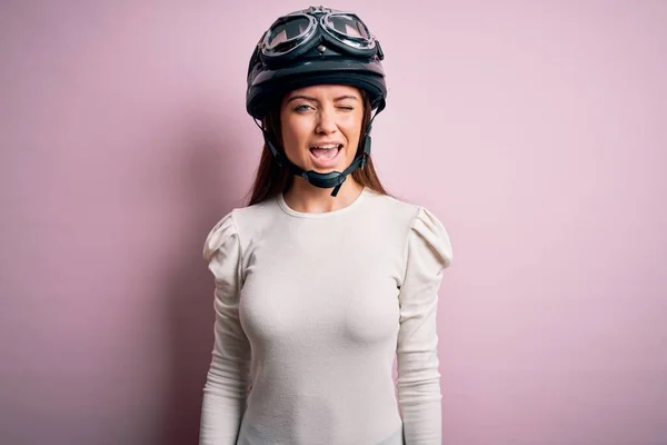 若いです美しいですオートバイの女性とともに青い目を身に着けています本ヘルメット上のピンクの背景ウィンクセクシーな表現でカメラを見ます 陽気で幸せな顔 — ストック写真