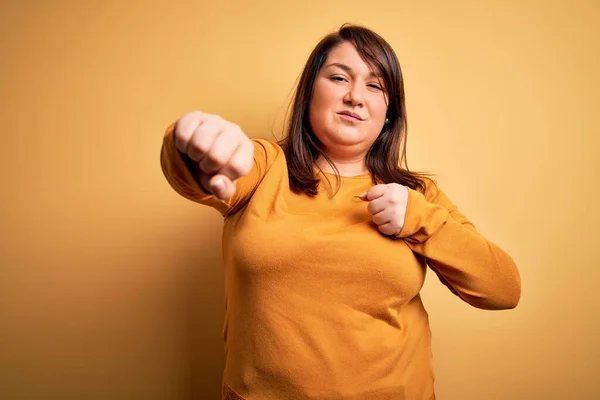 美しいブルネットプラスサイズ女性身に着けていますカジュアルセーター上の隔離された黄色の背景パンチ拳に戦い 積極的かつ怒りの攻撃 脅威と暴力 — ストック写真