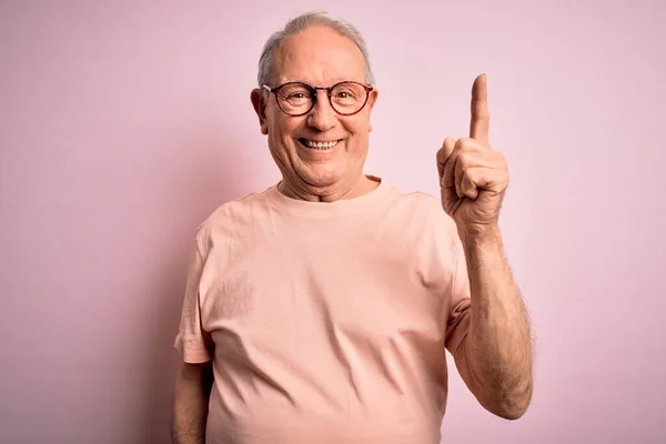 Grey haired senior man wearing glasses standing over pink isolated background pointing finger up with successful idea. Exited and happy. Number one.