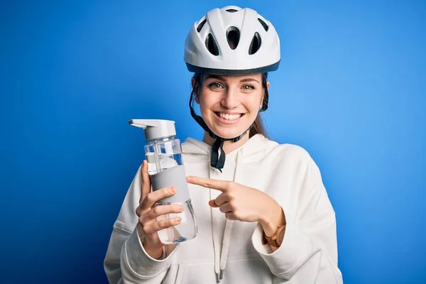 Young beautiful redhead cyclist woman wearing bike helmet drinkjng bottle of water very happy pointing with hand and finger